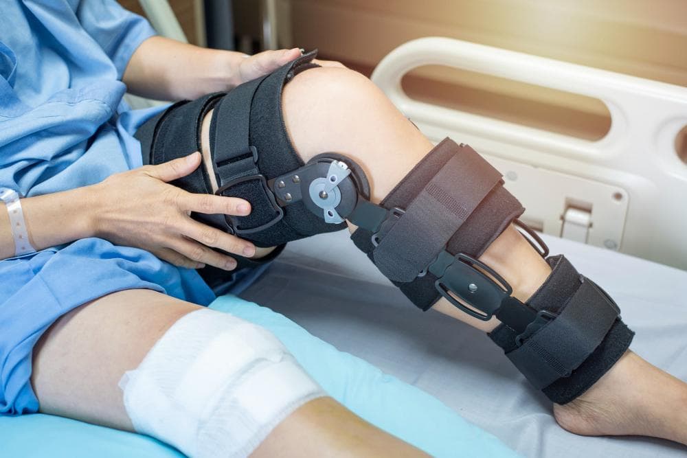 A patient is lying down in the hospital room post-surgery and is wearing a knee brace for proper recovery.