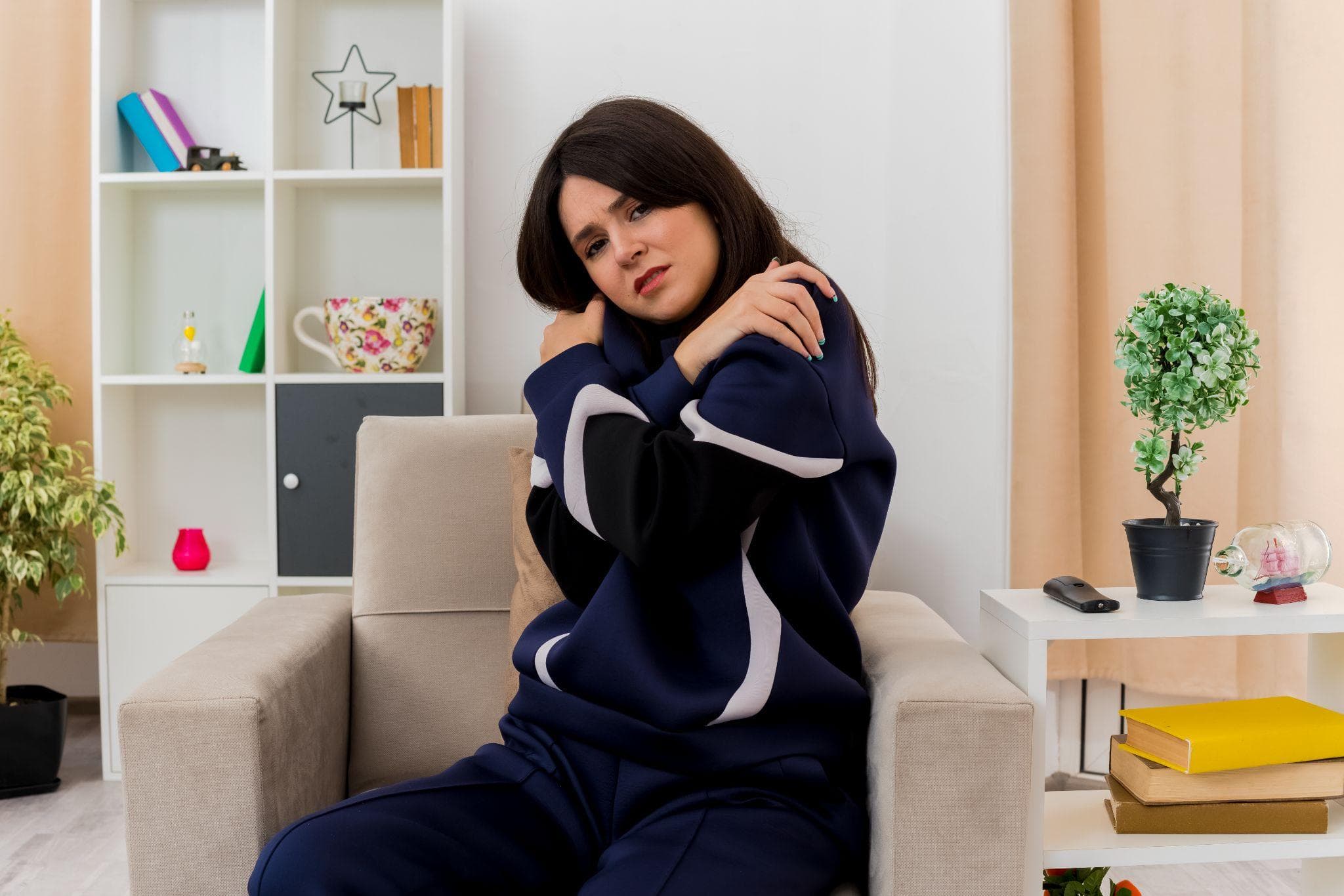 Young woman sitting on a chair holding her shoulders due to cold or pain.