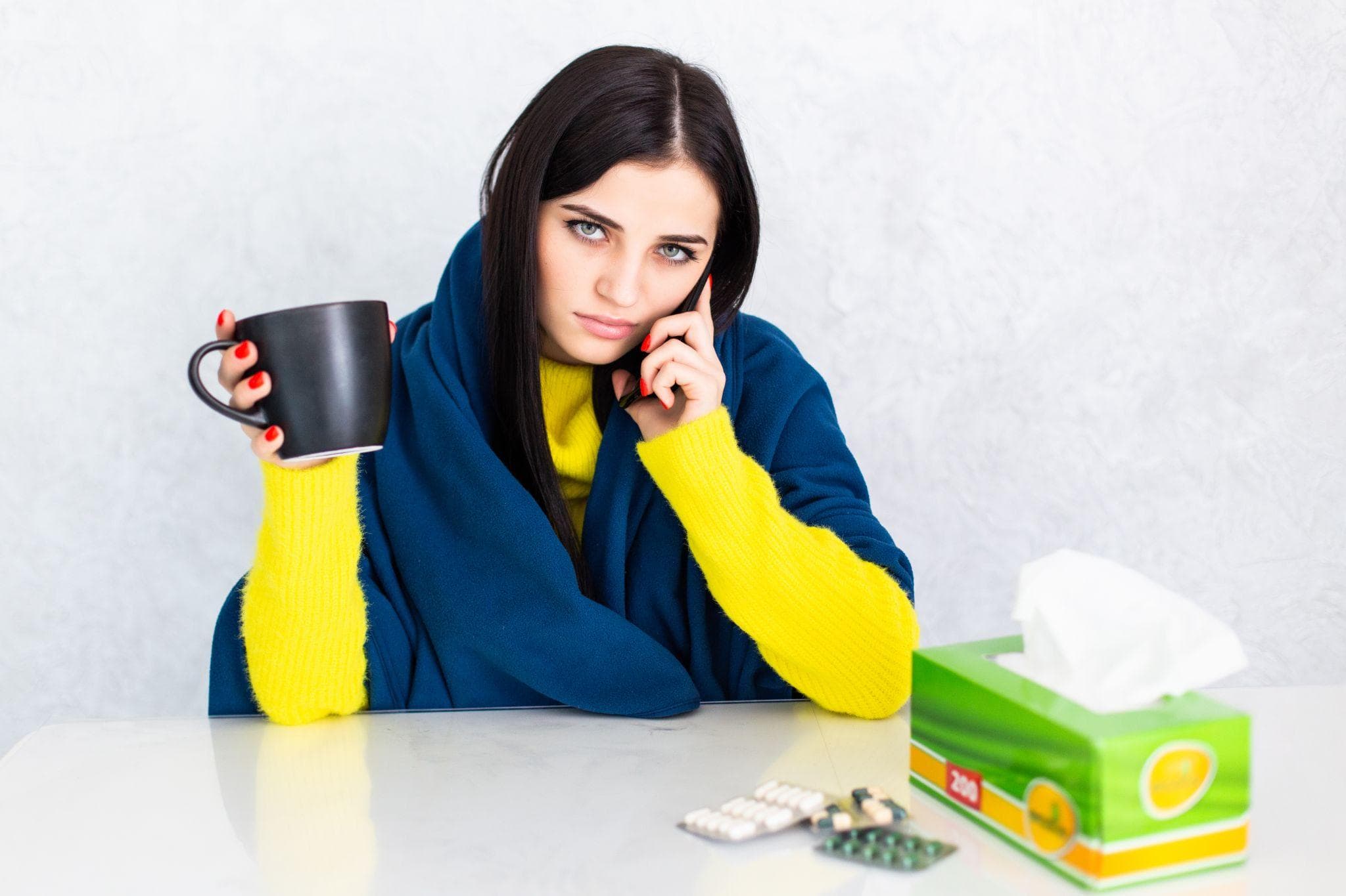 Sick woman wrapped in a blanket holding a cup of tea at the table.
