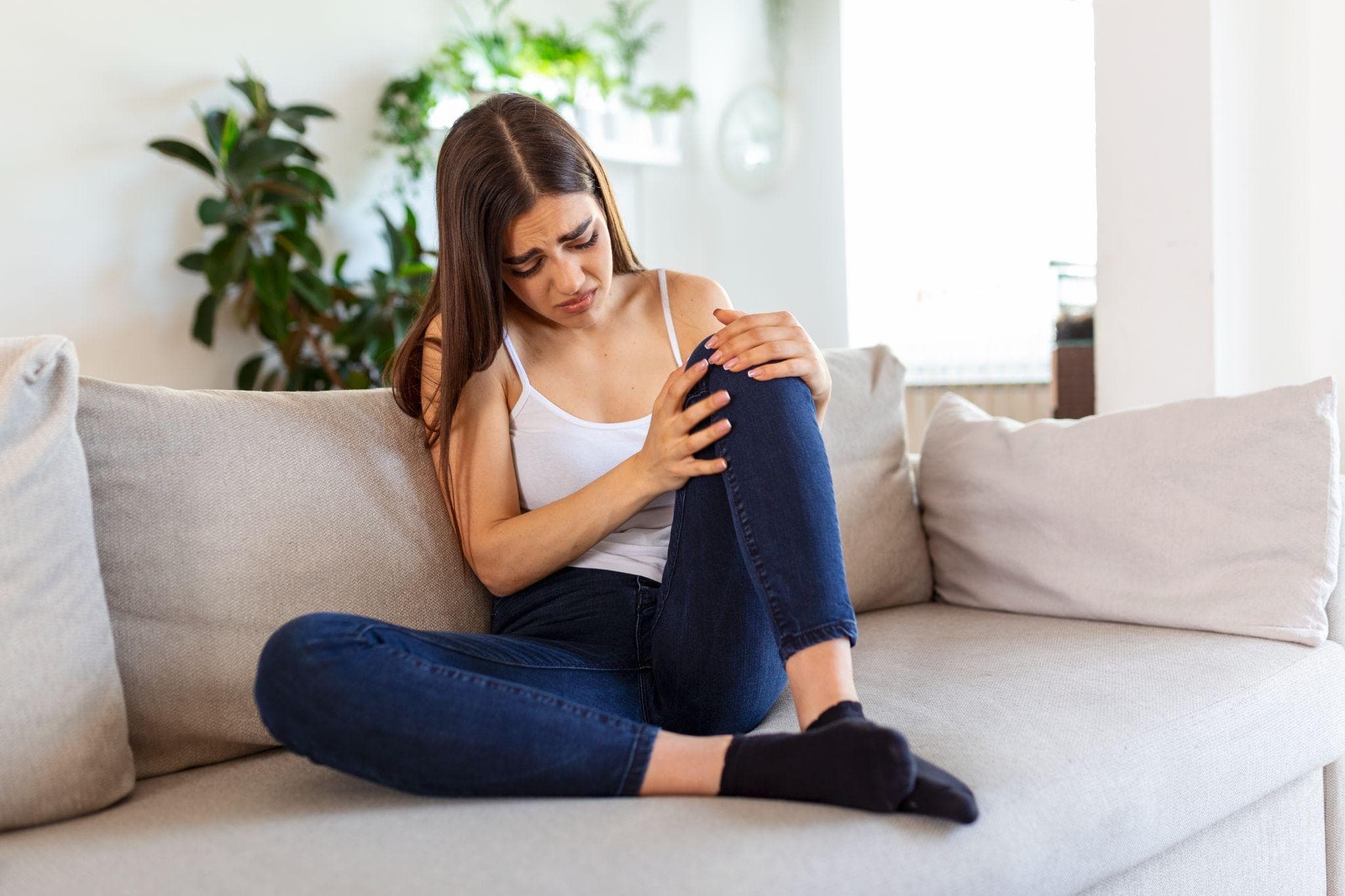 A young woman sitting on a beige sofa at home, wearing a white tank top and dark blue jeans, holding her knee with both hands and showing a painful expression on her face, indicating possible knee pain or discomfort.