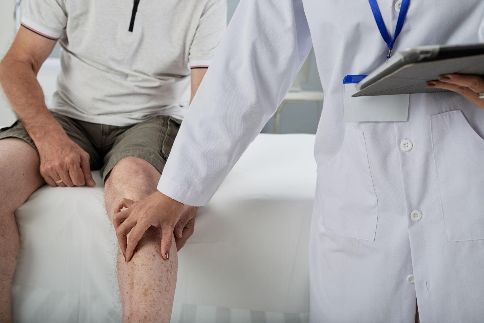 A male patient wearing a gray polo shirt and khaki shorts sitting on a medical examination table while a doctor in a white coat examines his knee and lower leg. The doctor is gently pressing on the patient’s shin, possibly assessing pain or swelling, while holding a clipboard for notes.