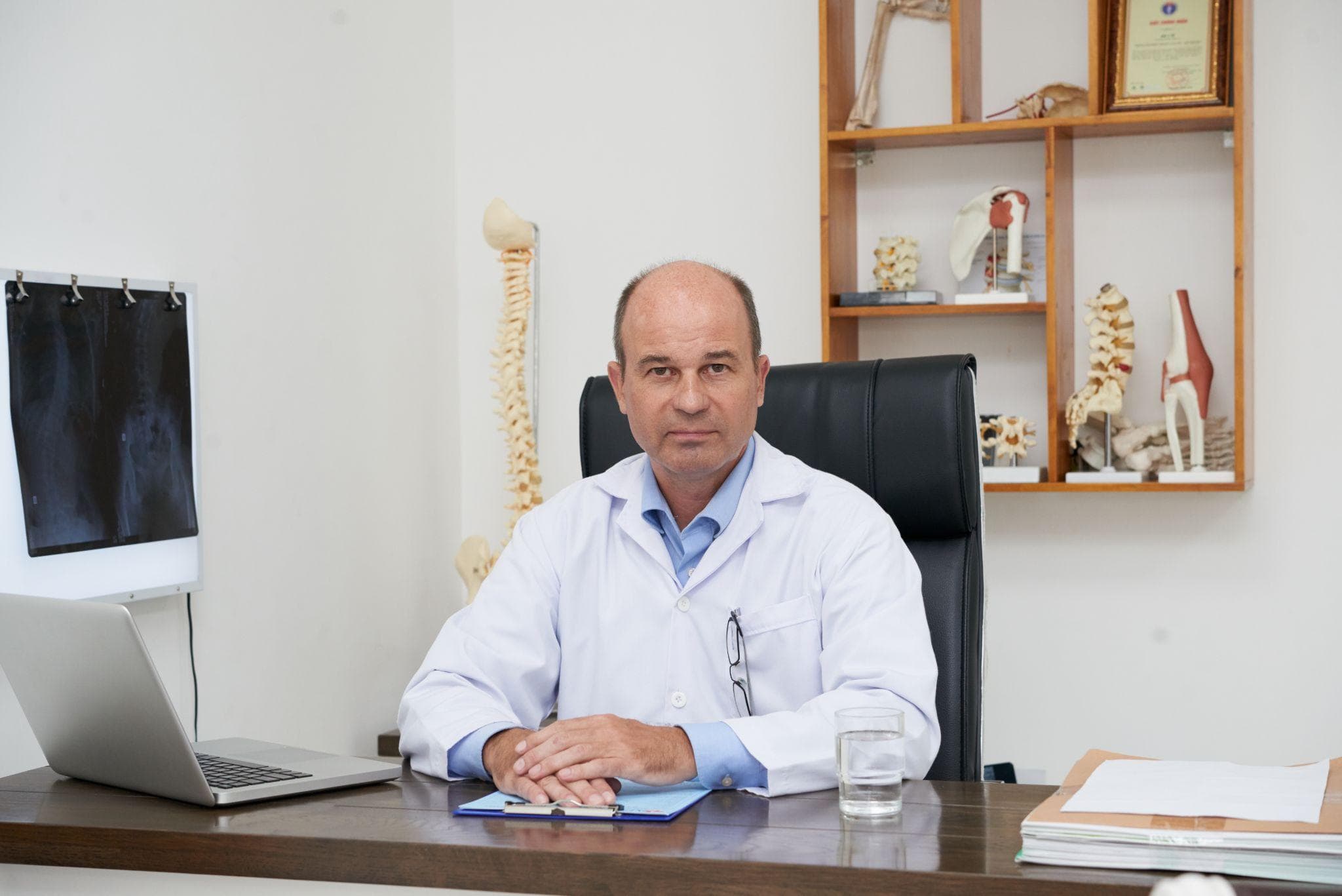 Middle-aged male orthopedic doctor sitting at his clinic desk with a laptop, medical reports, and spine models in the background.