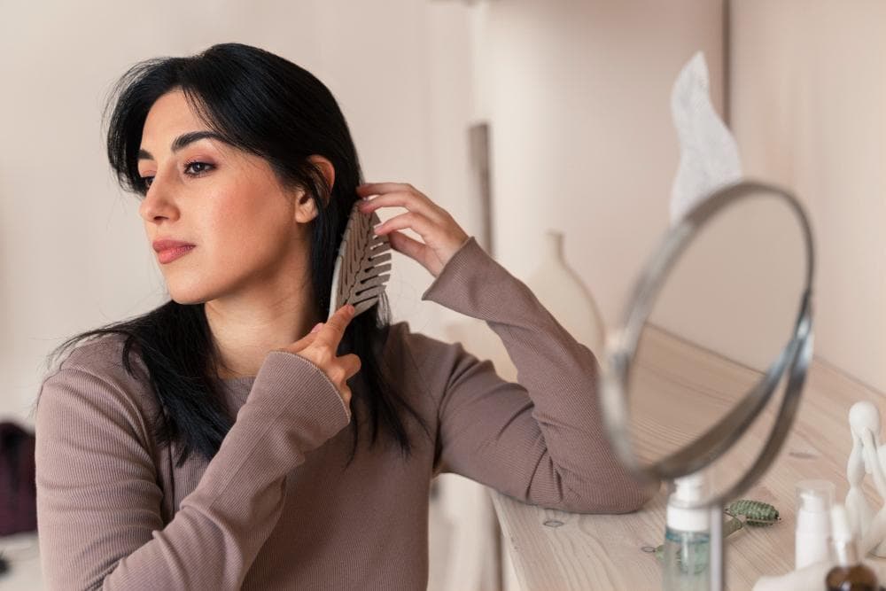 A woman is combing her hair in front of a short portable mirror in her room, and her skin products are kept on a table.