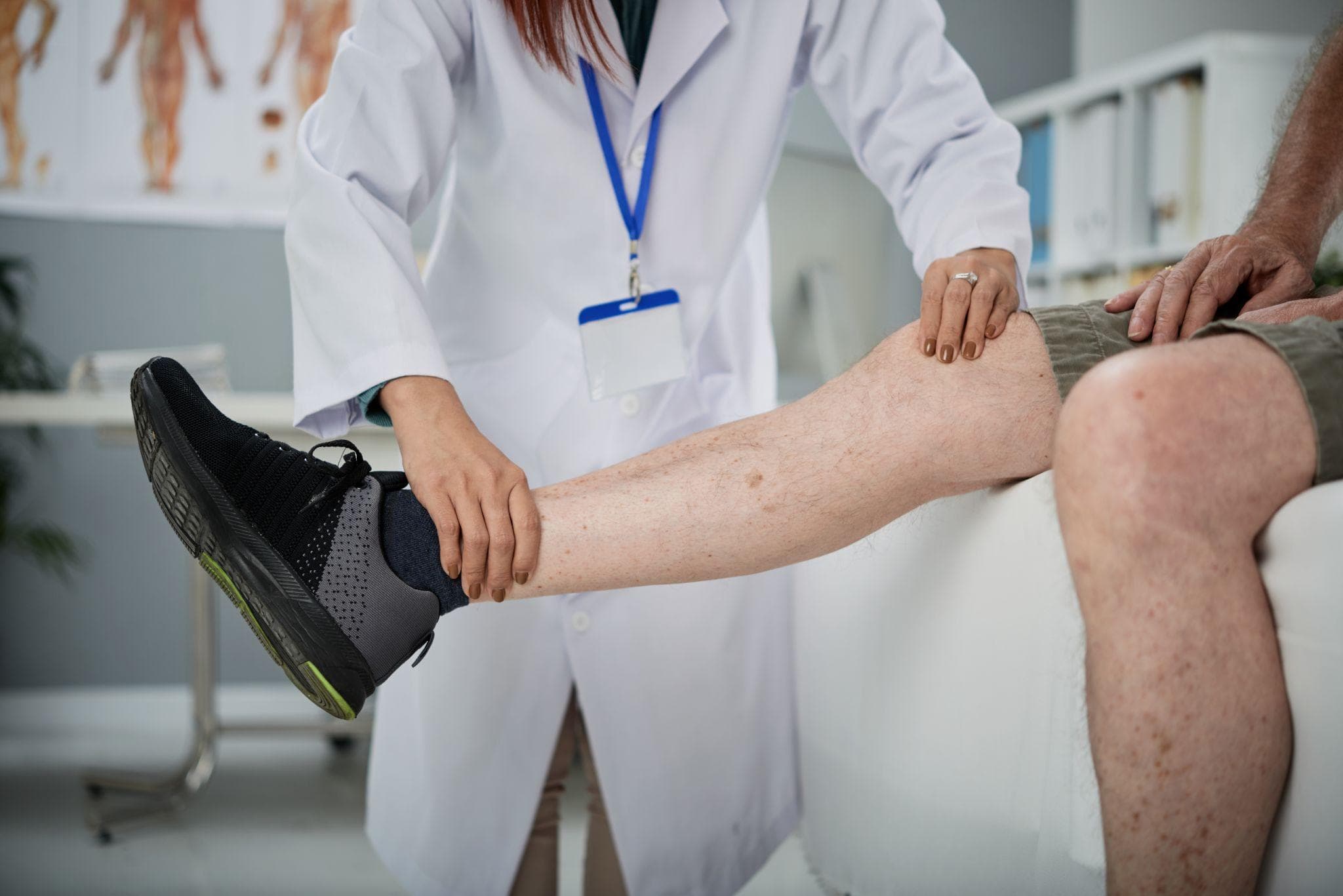 Female healthcare professional in a white coat examining a male patient’s knee by holding and assessing his lower leg in a clinical setting.