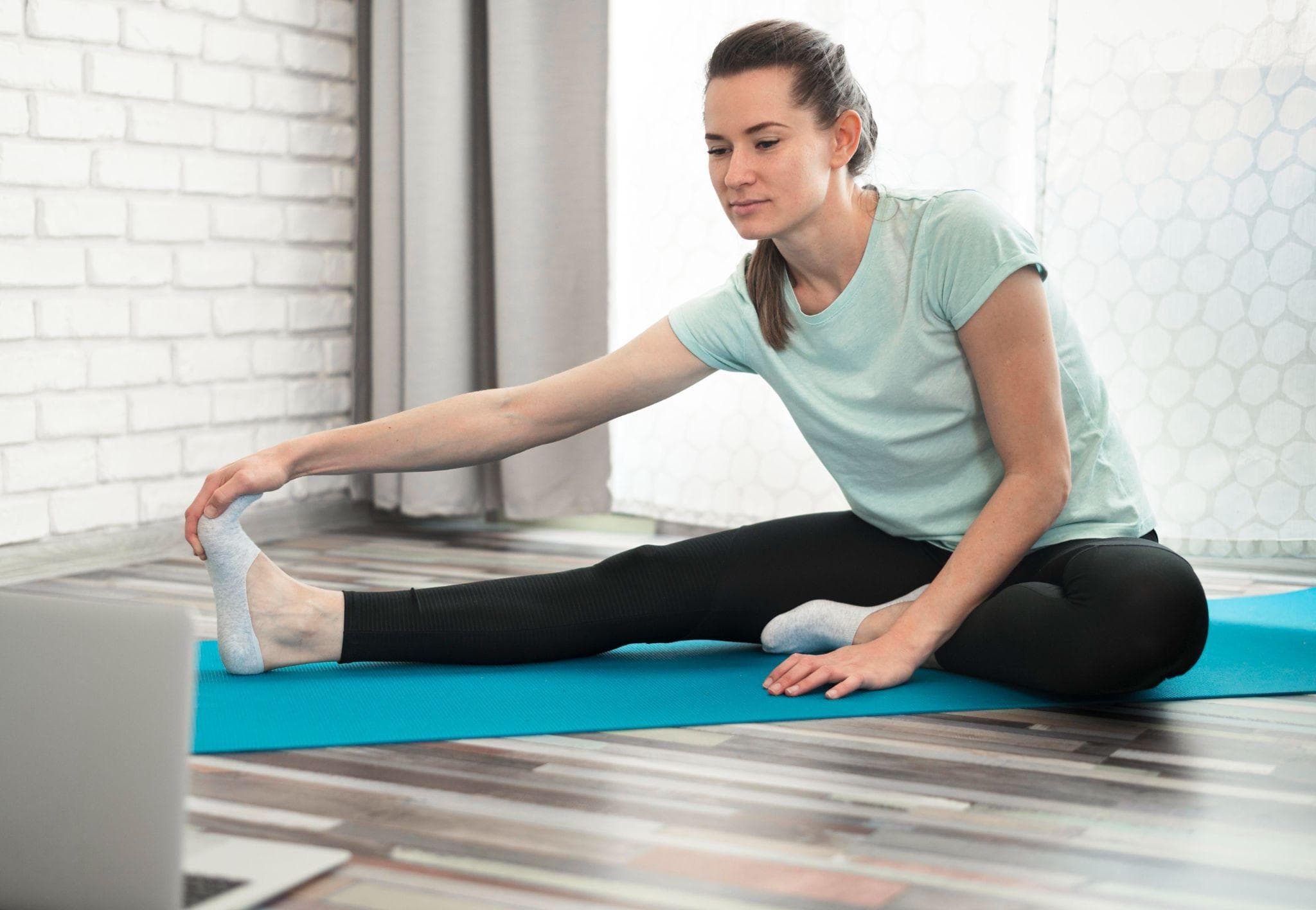 Young woman performing a seated hamstring stretch on a yoga mat at home, reaching toward her extended foot during a morning workout session.