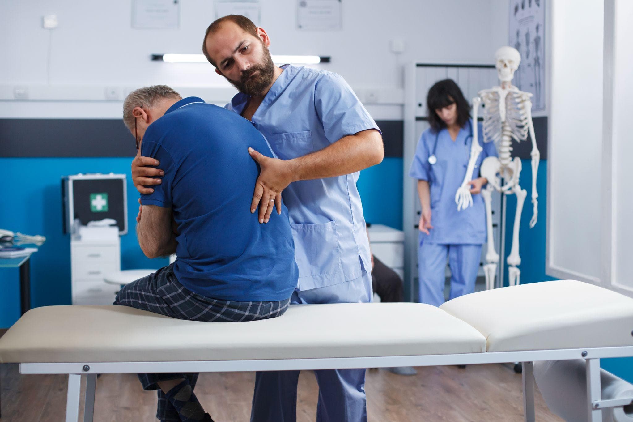 A male physiotherapist is providing manual therapy to a middle-aged man sitting on a treatment table in a clinic. At the same time, another medical staff member and a skeleton model are visible in the background.
