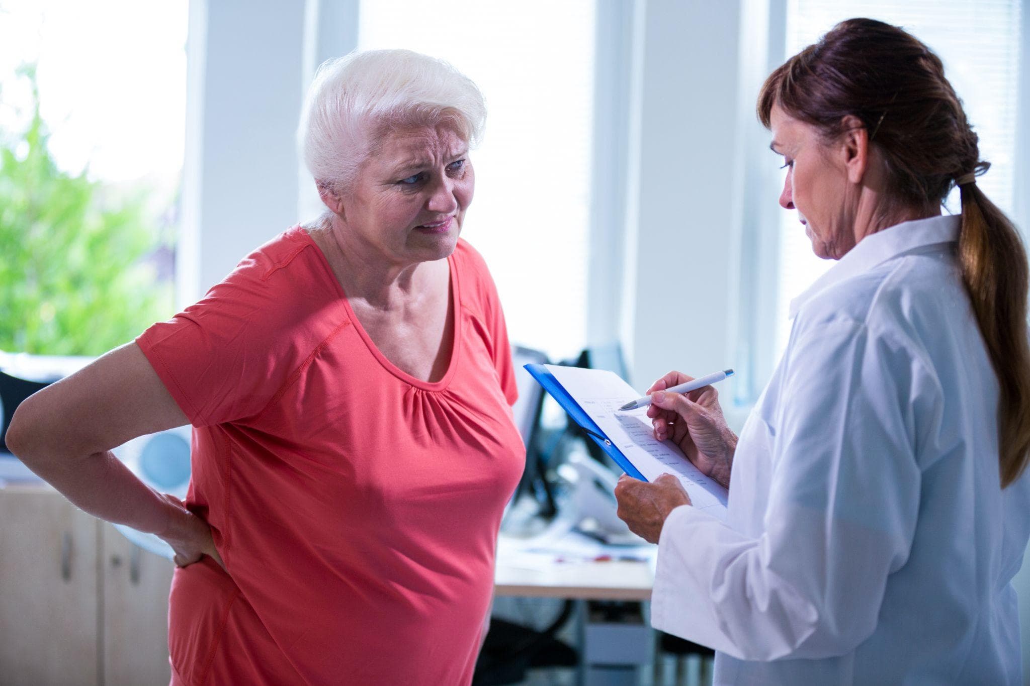 A female doctor is making notes and examining an elderly female patient as she stands in front of her, holding her back in pain.
