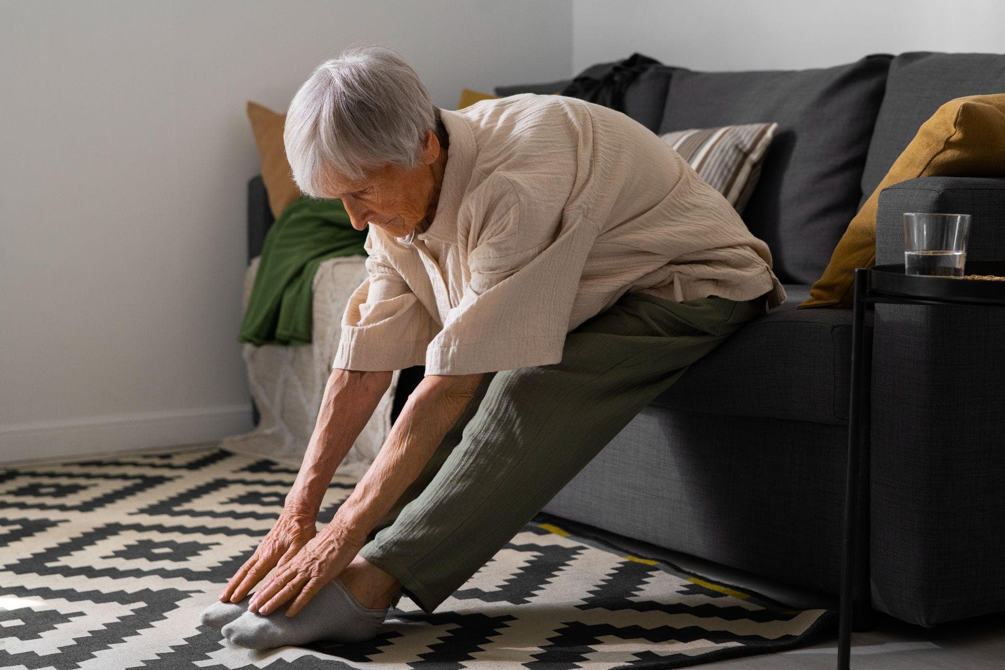 Senior woman sitting on a sofa at home, bending forward to stretch her legs and touch her toes as part of a light exercise routine in a living room environment.