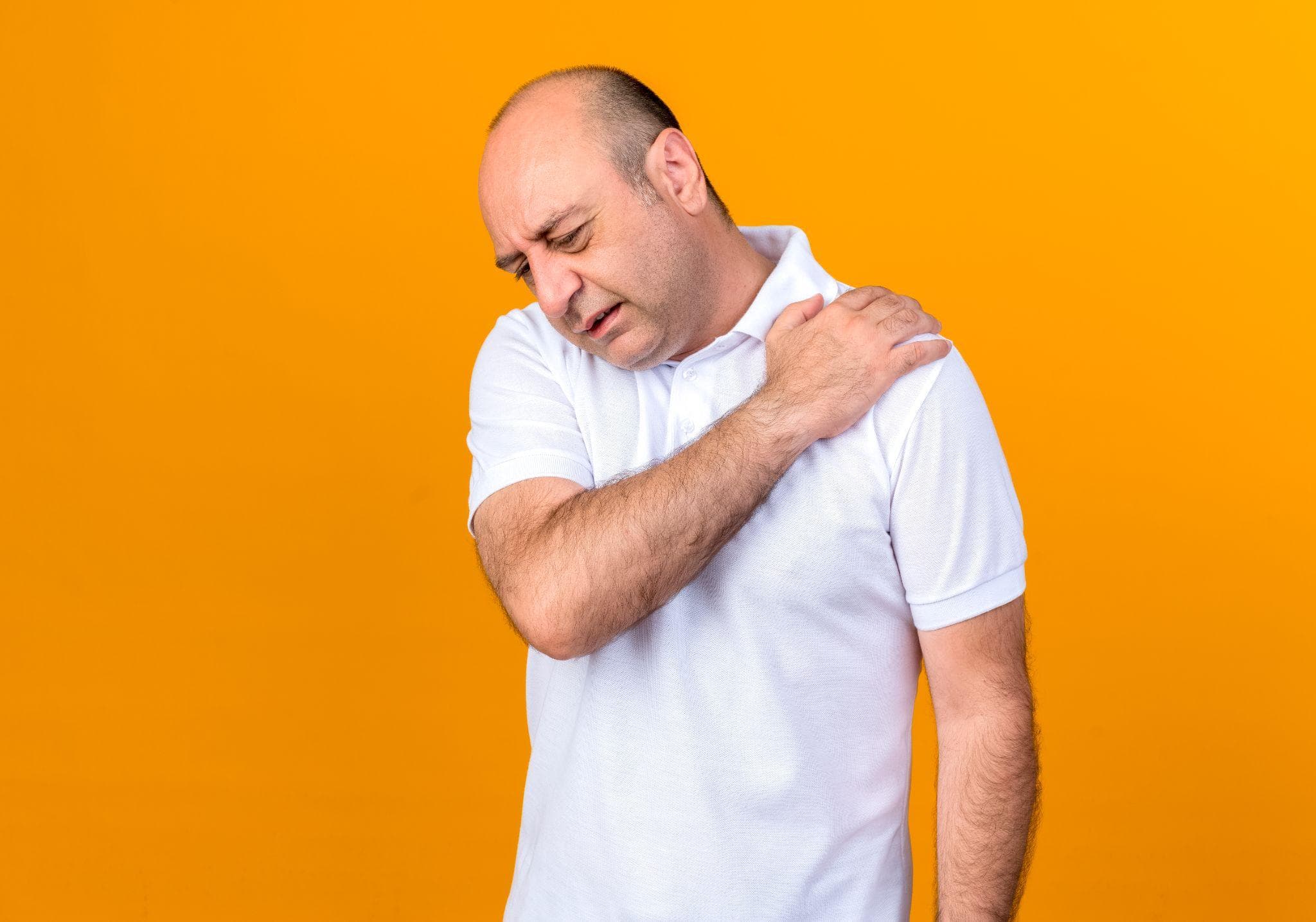 A man is standing against an orange background, holding his shoulder in pain.