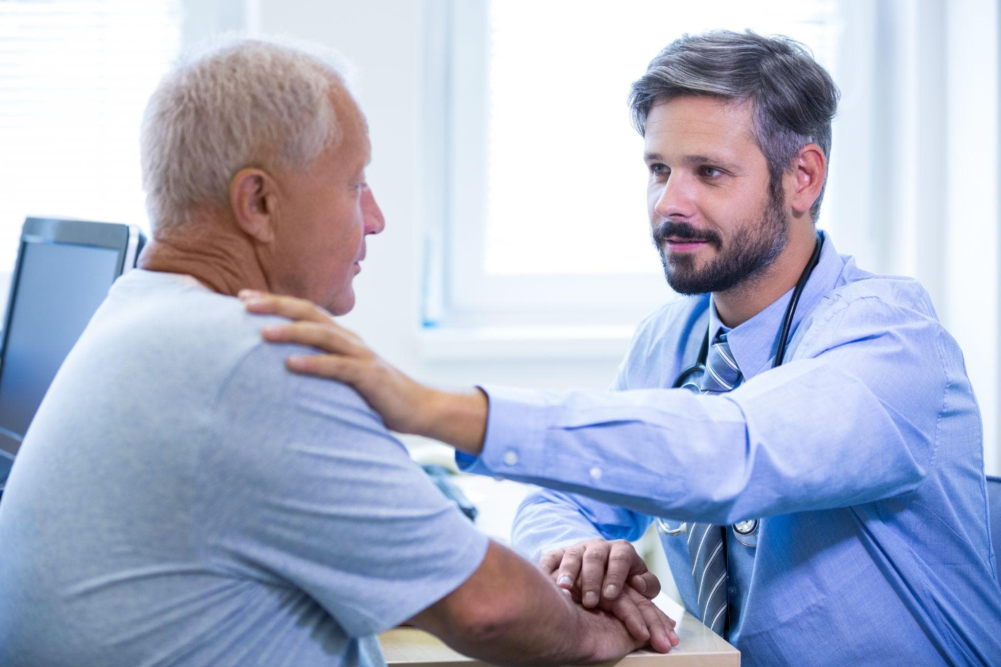 A doctor is examining the shoulder of an elderly patient in his clinic with an empathetic look on his face.