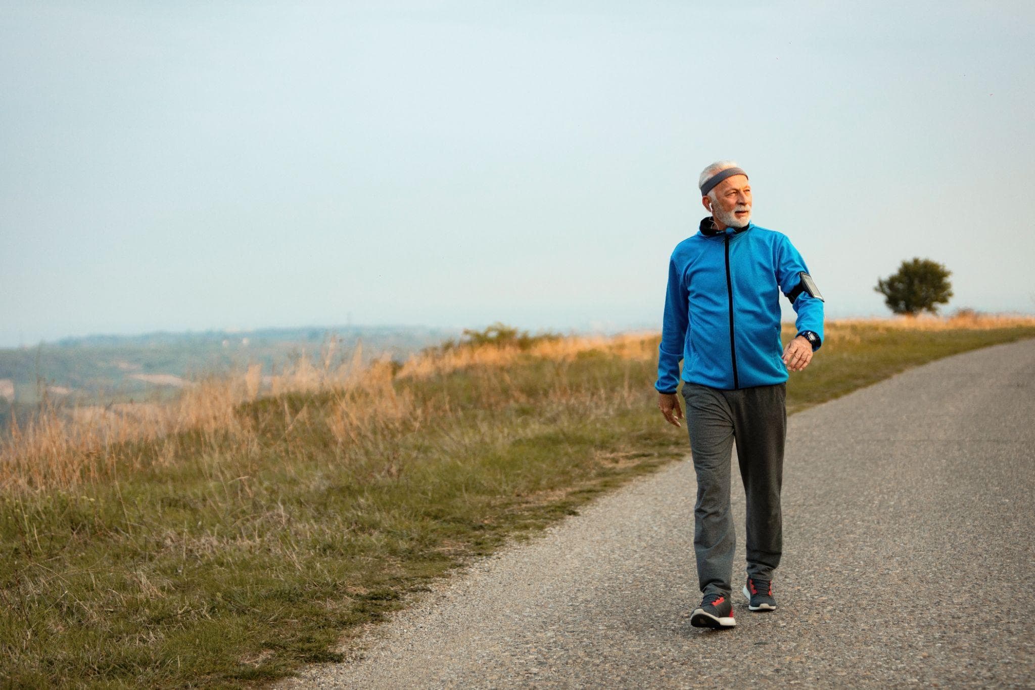 Full-length senior man in sportswear walking outdoors on a countryside road during morning hours, wearing earphones and a fitness band, staying active and maintaining a healthy lifestyle.