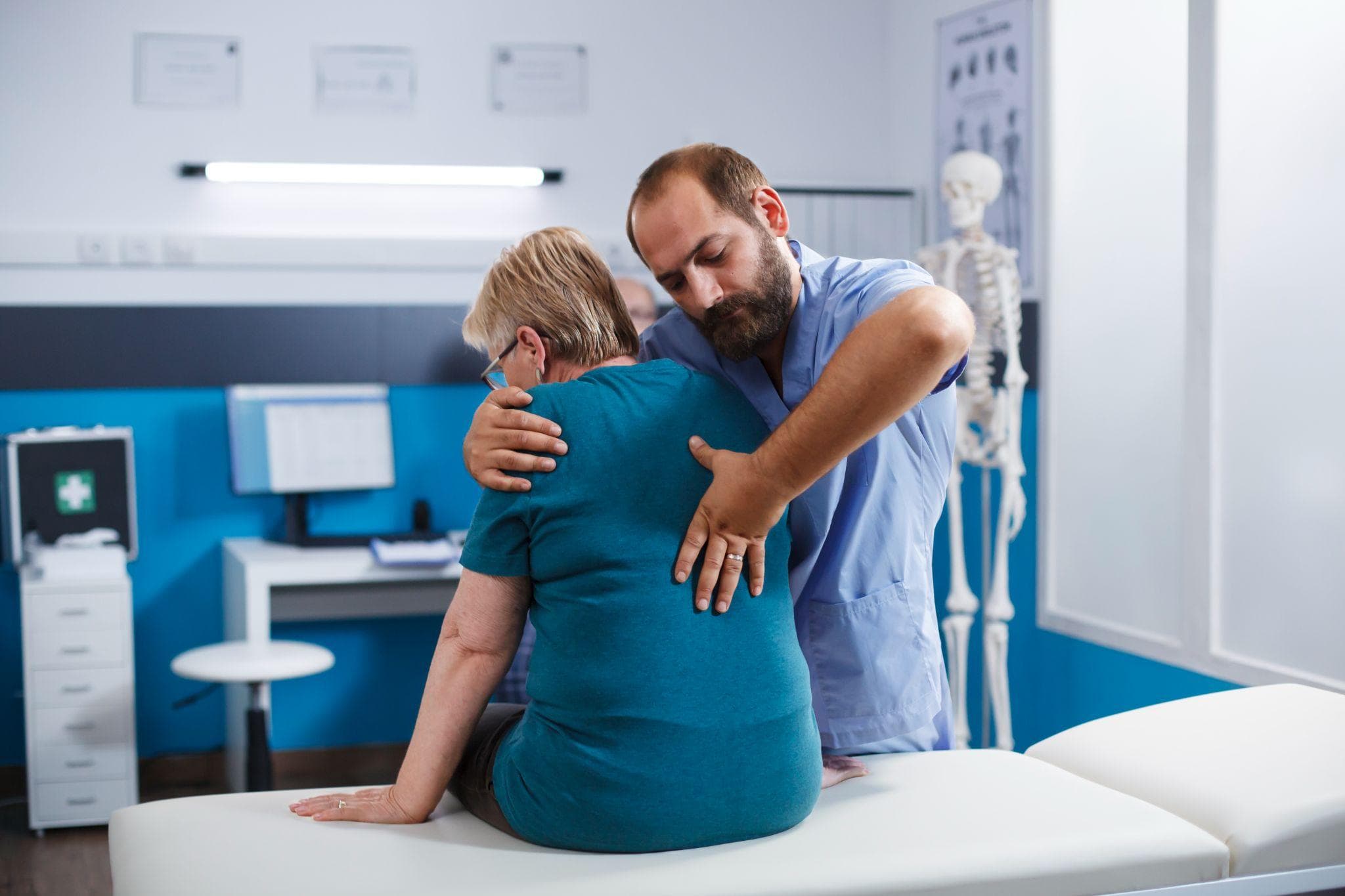 A male physiotherapist is examining and applying manual therapy on an elderly woman’s upper back while she sits on an examination table in a medical clinic, with a human skeleton model visible in the background.