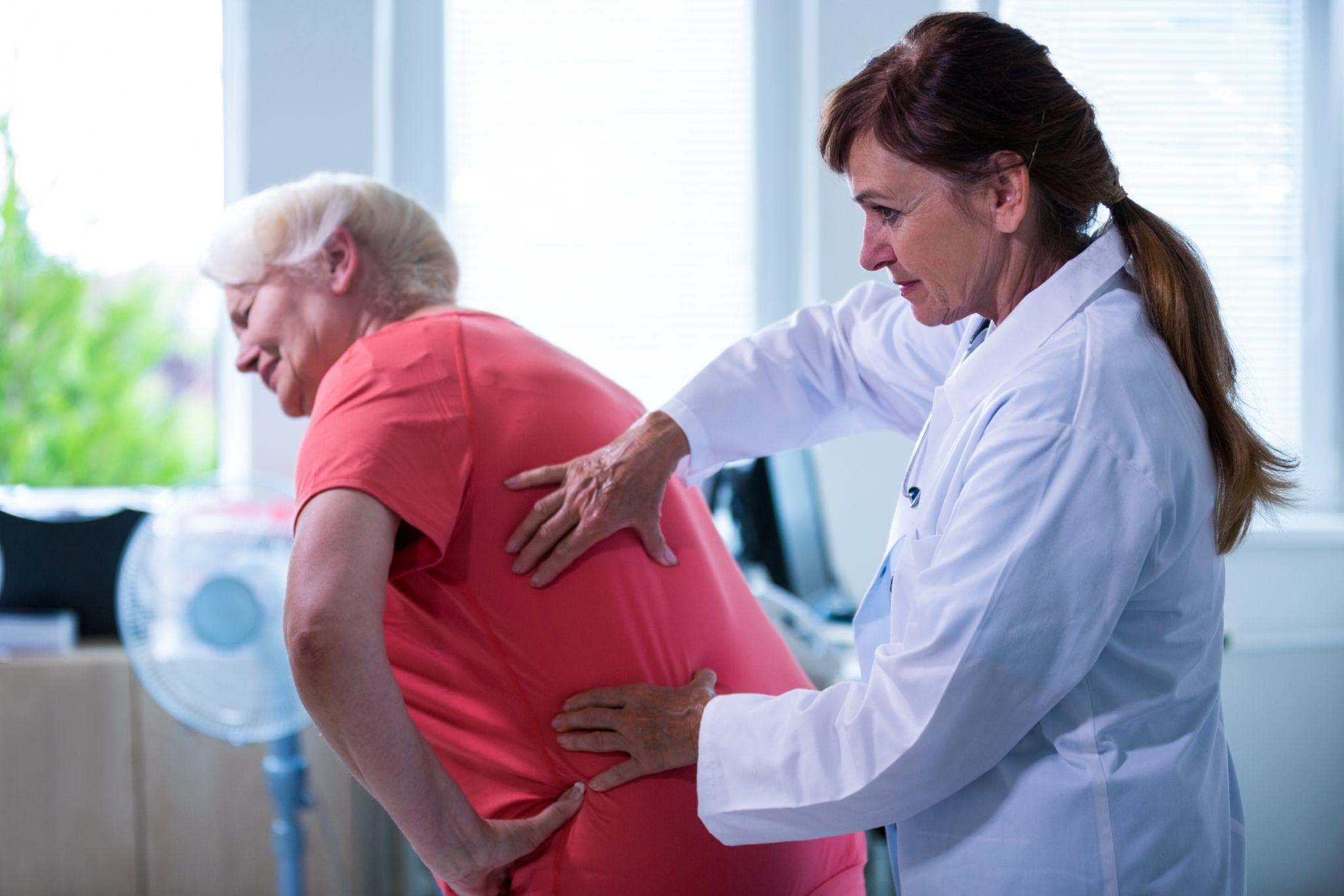 A female doctor is examining the back of the elderly patient, who seems in visible pain, in her clinic.