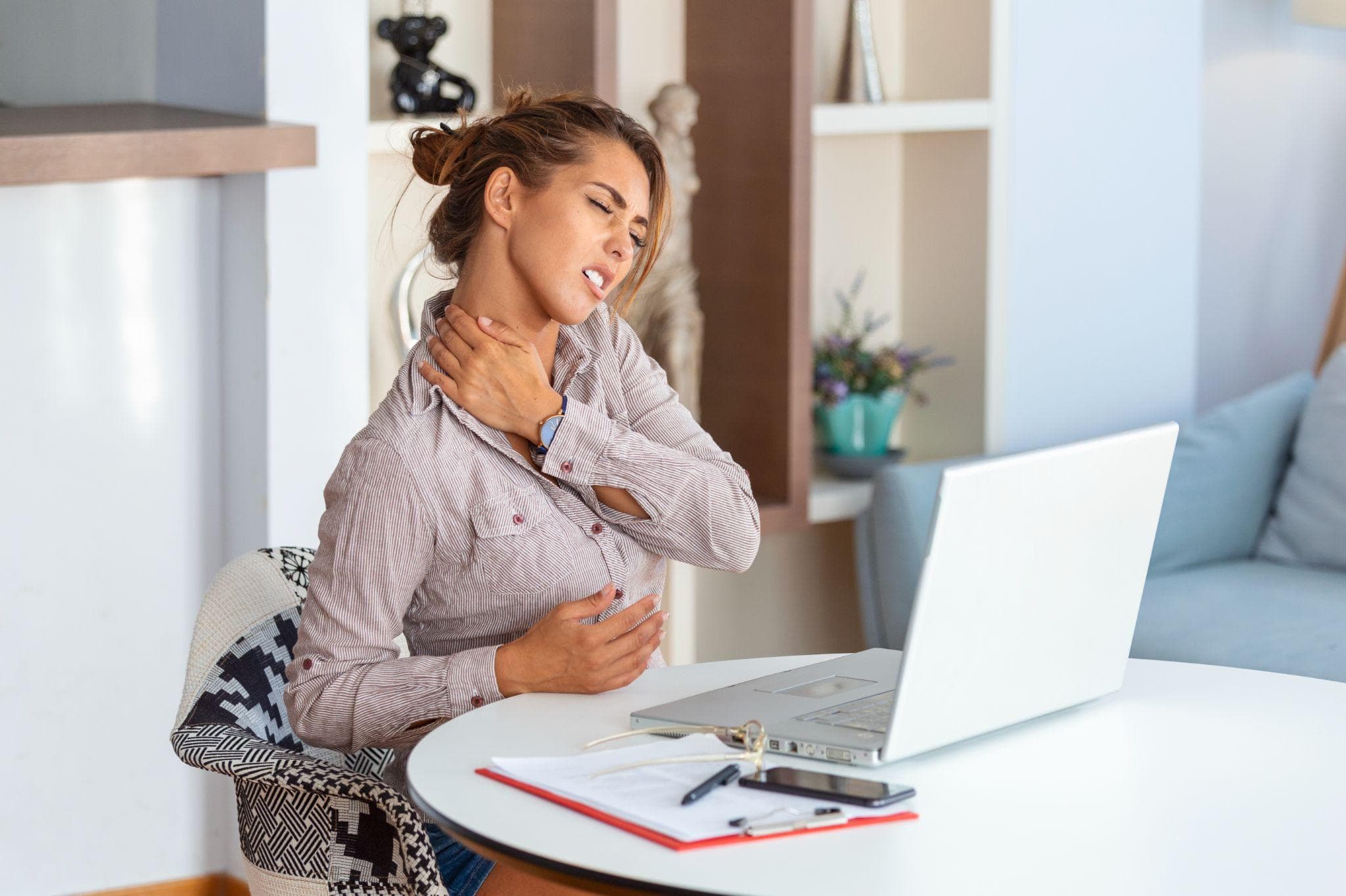 Young businesswoman sitting at a desk in a home office, holding her neck in pain while looking at her laptop screen.