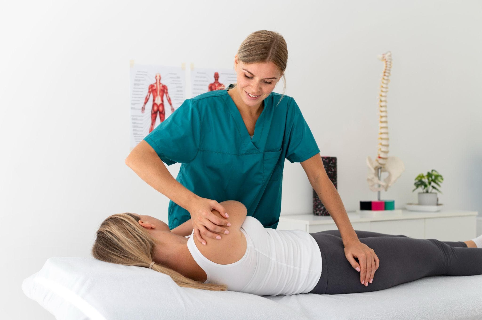Female physiotherapist in green medical scrubs performing shoulder mobility therapy on a woman lying on a treatment table in a clinical setting, with a spine model and anatomical posters visible in the background.