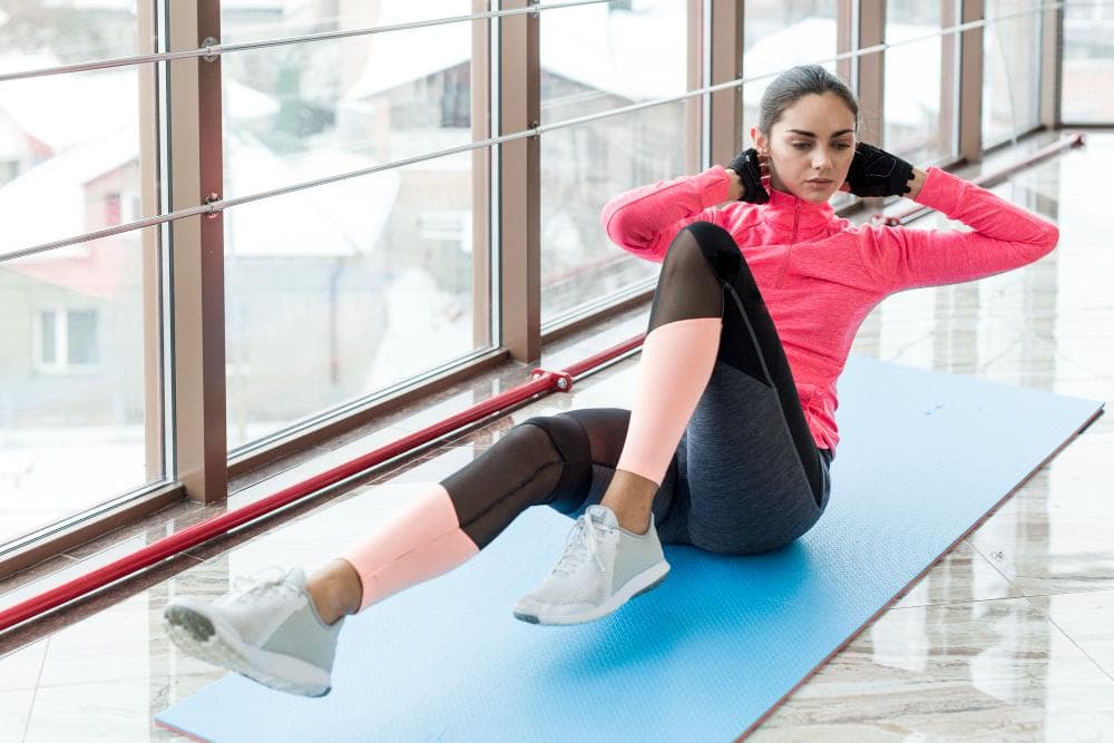 A woman is exercising on a blue mat in her room near the windows.