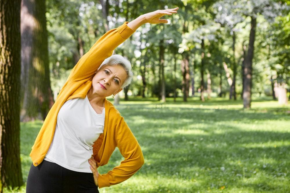 A senior woman doing side stretching exercise outdoors in a park to maintain bone strength and flexibility after menopause.