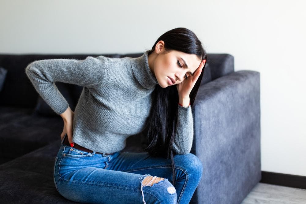 A woman is sitting on her couch in her home, bending down in pain while holding her lower back.