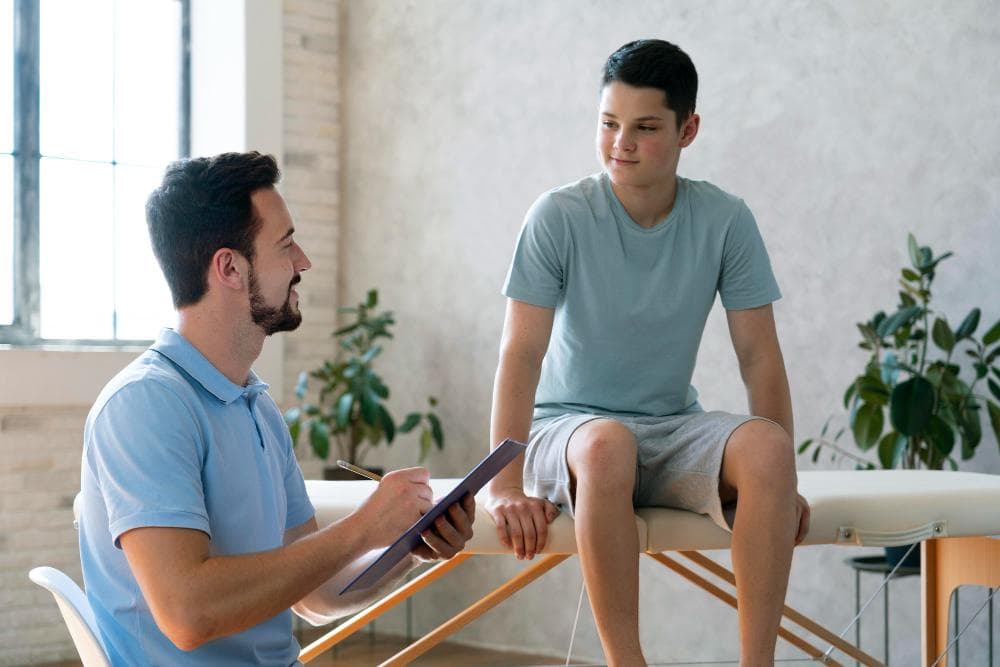 Physiotherapist consulting a teenage boy sitting on an examination table in a clinic, assessing knee or leg pain while noting observations on a clipboard.