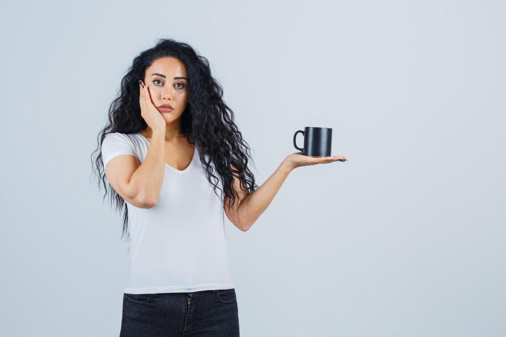 A woman is standing against a white background while holding a coffee mug with a confused expression on her face.