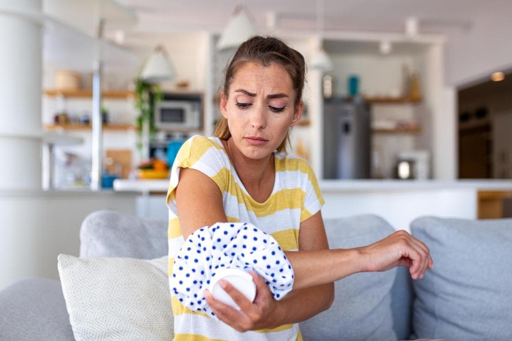 A young woman applying an ice pack to her elbow at home, showing discomfort and pain relief treatment commonly used for tennis elbow or muscle strain.