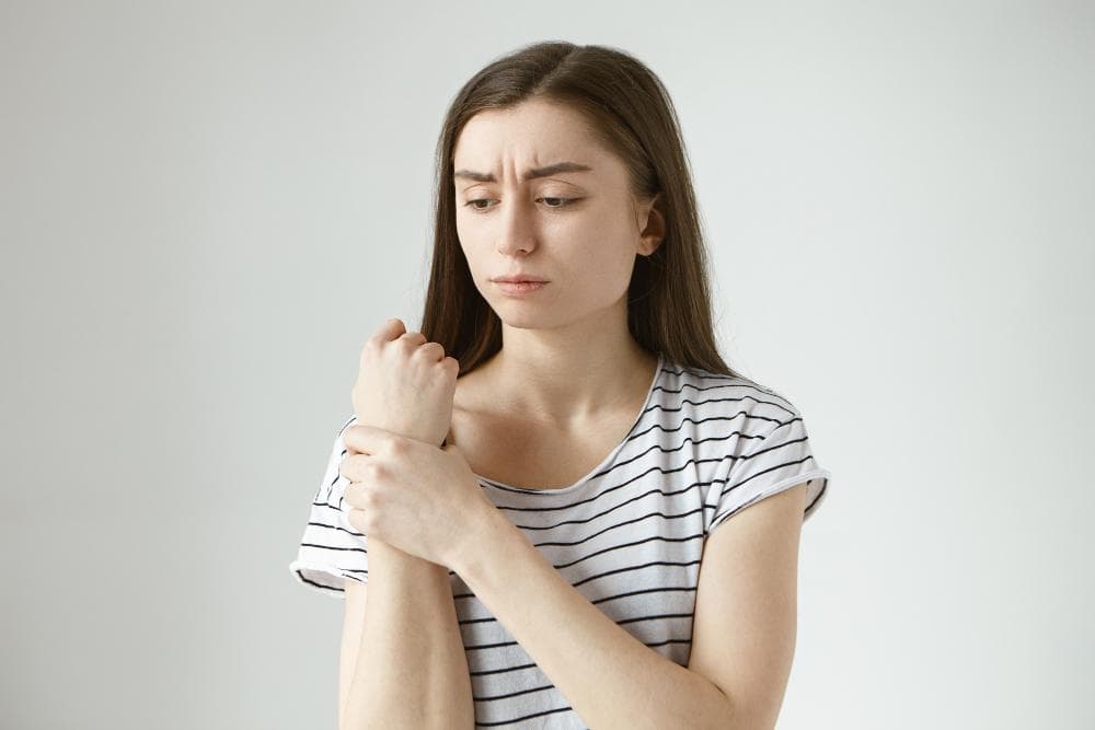 A woman in a striped top holds her wrist with one hand, indicating discomfort while looking at her other hand. The focus is on the wrist area, suggesting pain or injury.