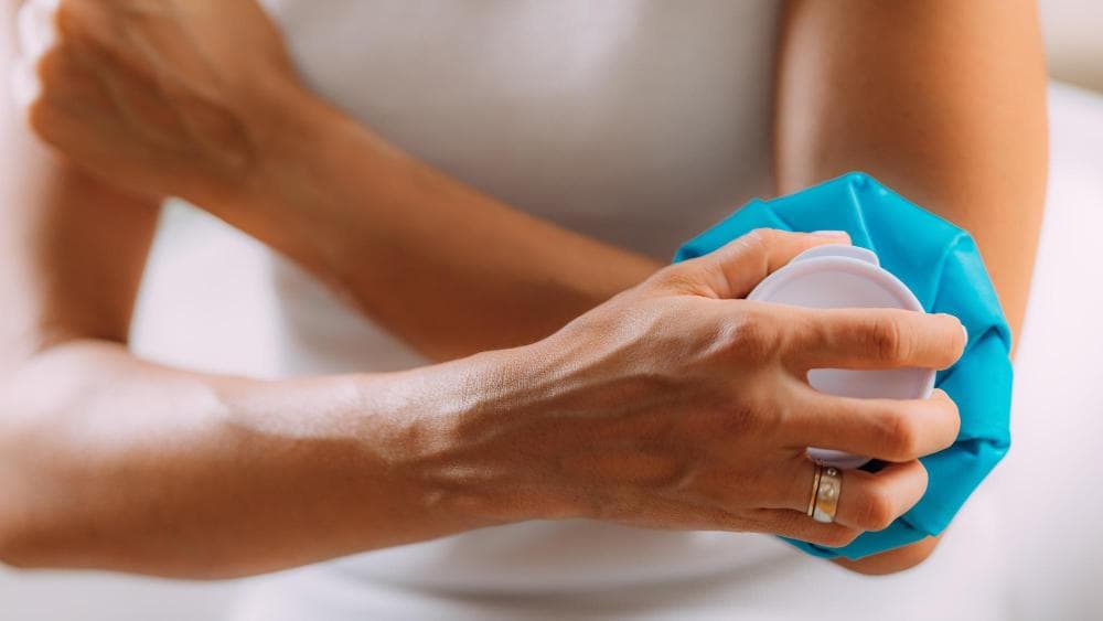 A close-up of a hand in a white shirt pressing a blue, moldable ice pack onto an elbow, showing the pack's contours around the joint.