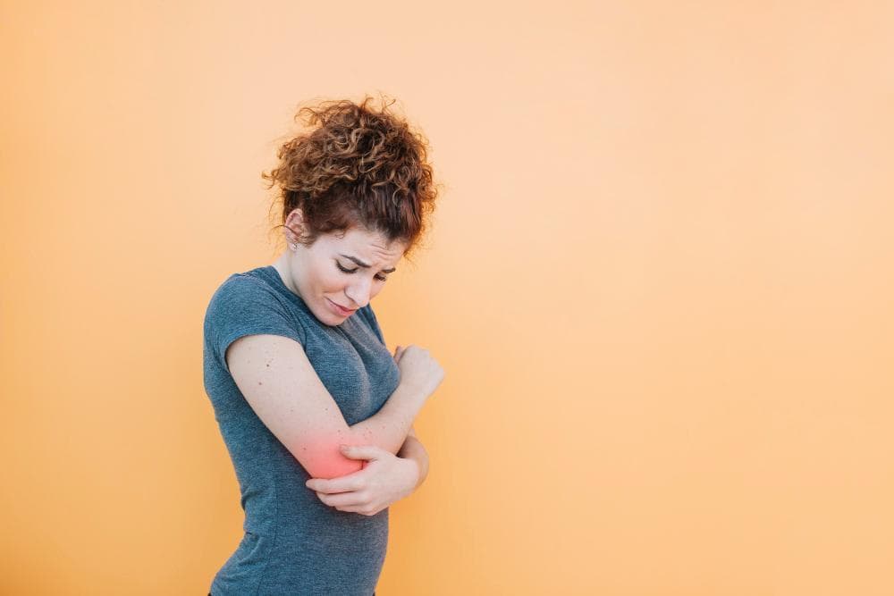 A young woman with curly hair wearing a gray t-shirt looks worried as she gently cradles her right elbow, set against a solid orange background.