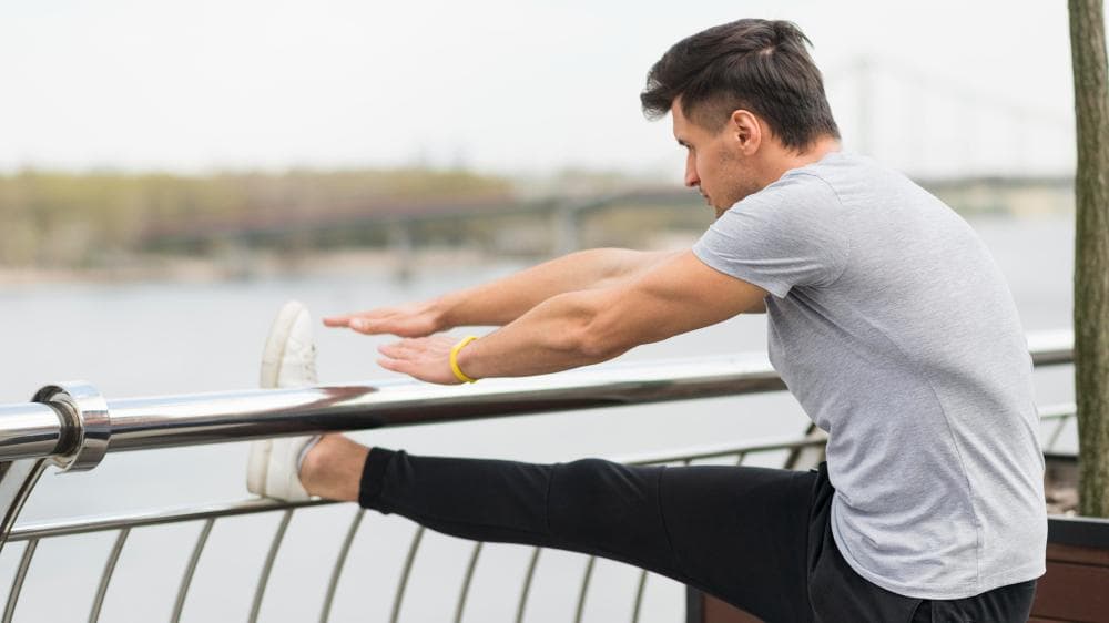 A man in an outside area is stretching his achilles tendon by putting his leg on a railing bar.