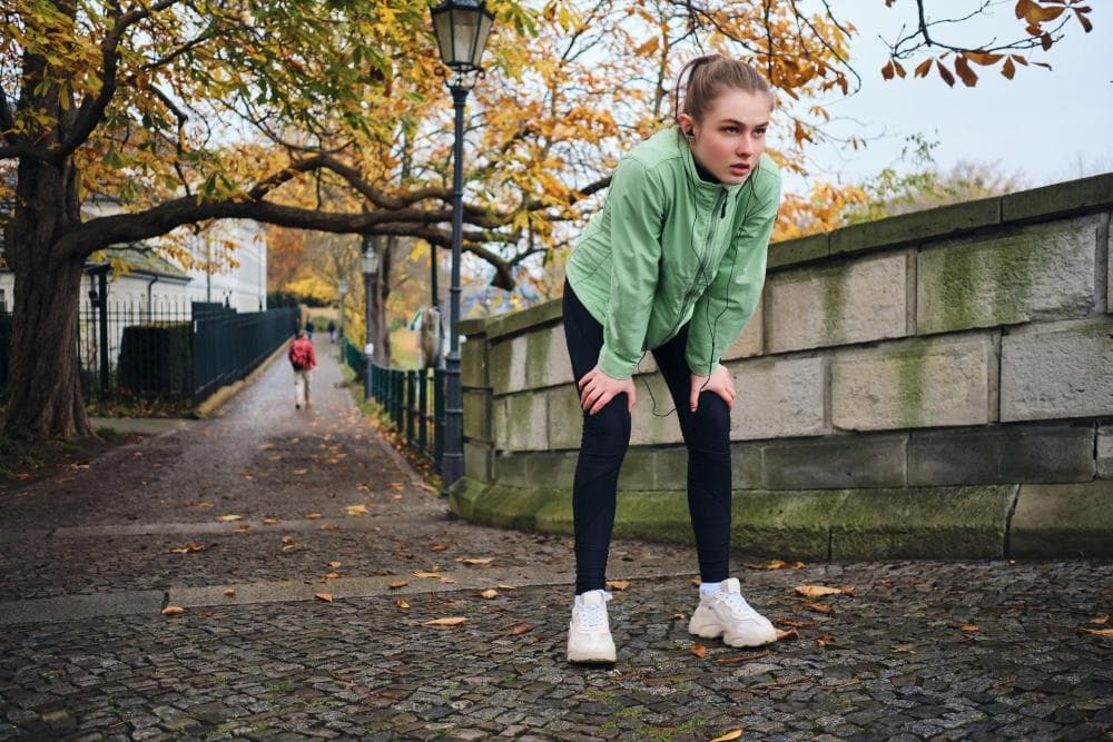 The young woman is bending forward with both hands placed on her knees and seems to be panting. She is outdoors. There are autumn leaves behind, and the ground has a cobblestone path and a lamppost in the background.