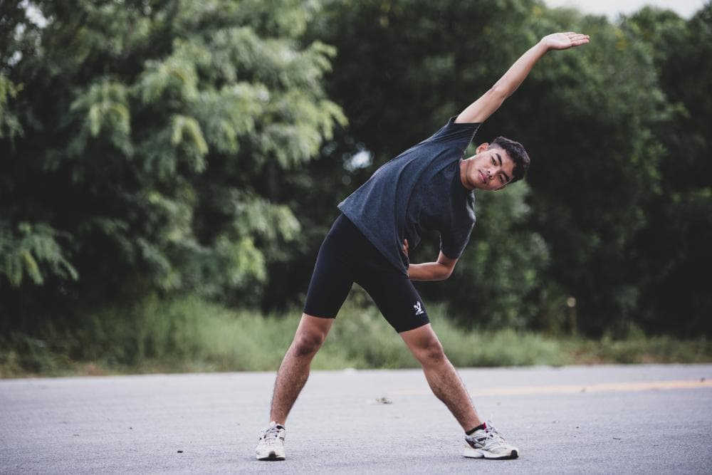 A young man is exercising outdoors by the side of a road. His face shows he is concentrated while raising his hand up in the air to stretch himself. There are lush green trees around as well in the background.