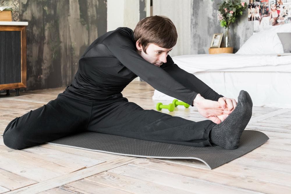 Young man performing a seated hamstring and hip stretch on a yoga mat indoors, reaching toward his foot while keeping one leg extended and the other bent, with dumbbells placed in the background.