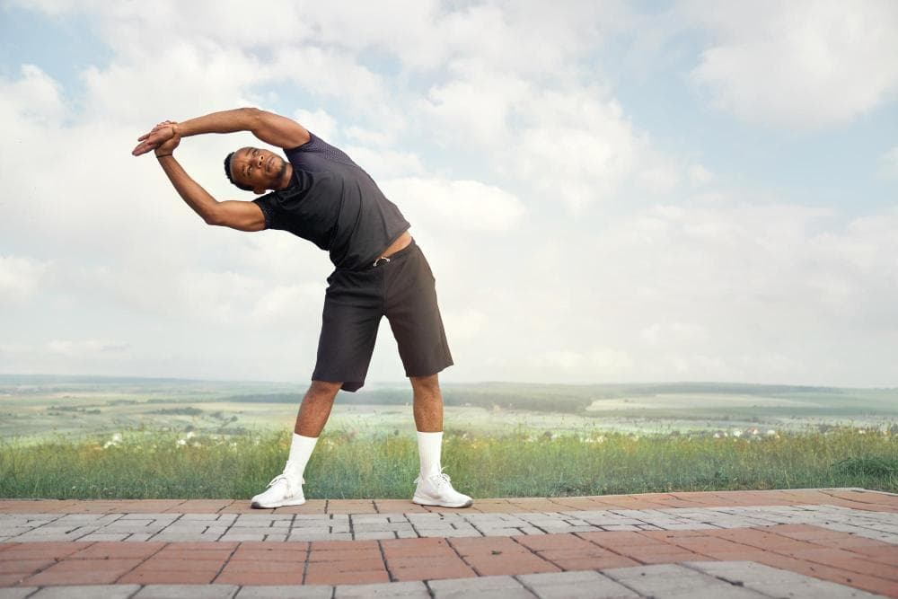 Young male athlete performing a side stretch exercise outdoors, standing on a paved surface with hands clasped overhead and body bent sideways, wearing sportswear with a scenic landscape and cloudy sky in the background.