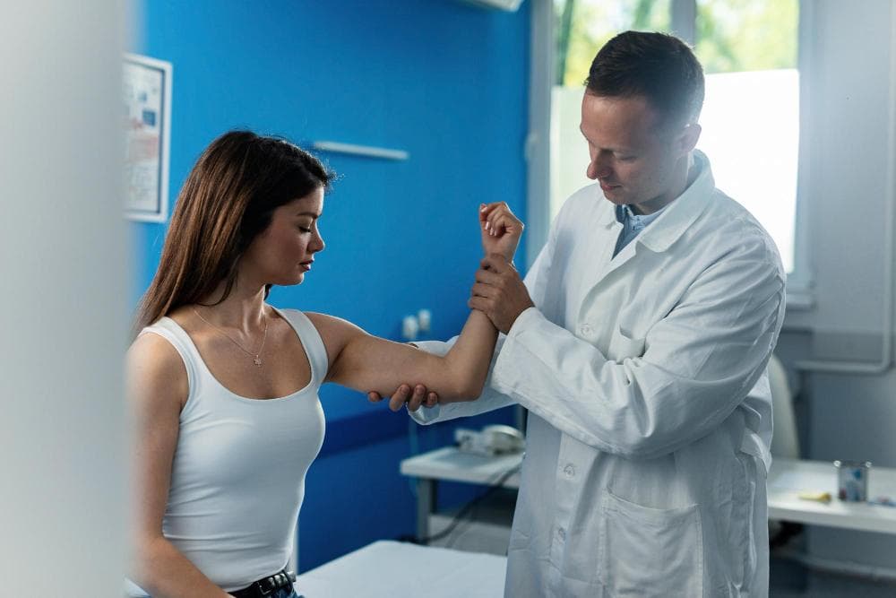 A female patient is being examined in a clinic by a doctor. The doctor is examining her arm muscles around the elbow.