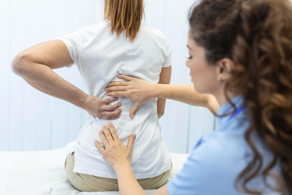 A female doctor is examining the back of the female patient in the clinic, and she seems to be in pain as she is holding her back.