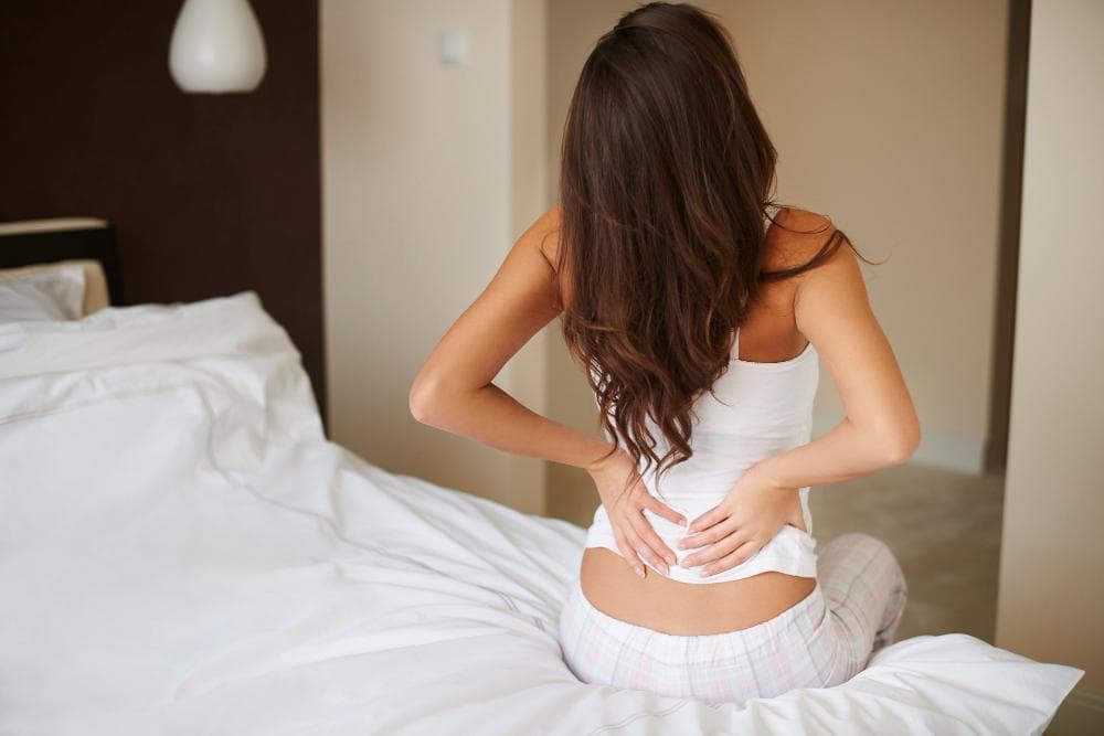 A woman’s back view can be seen sitting on her bed in her room, holding her lower back in pain.