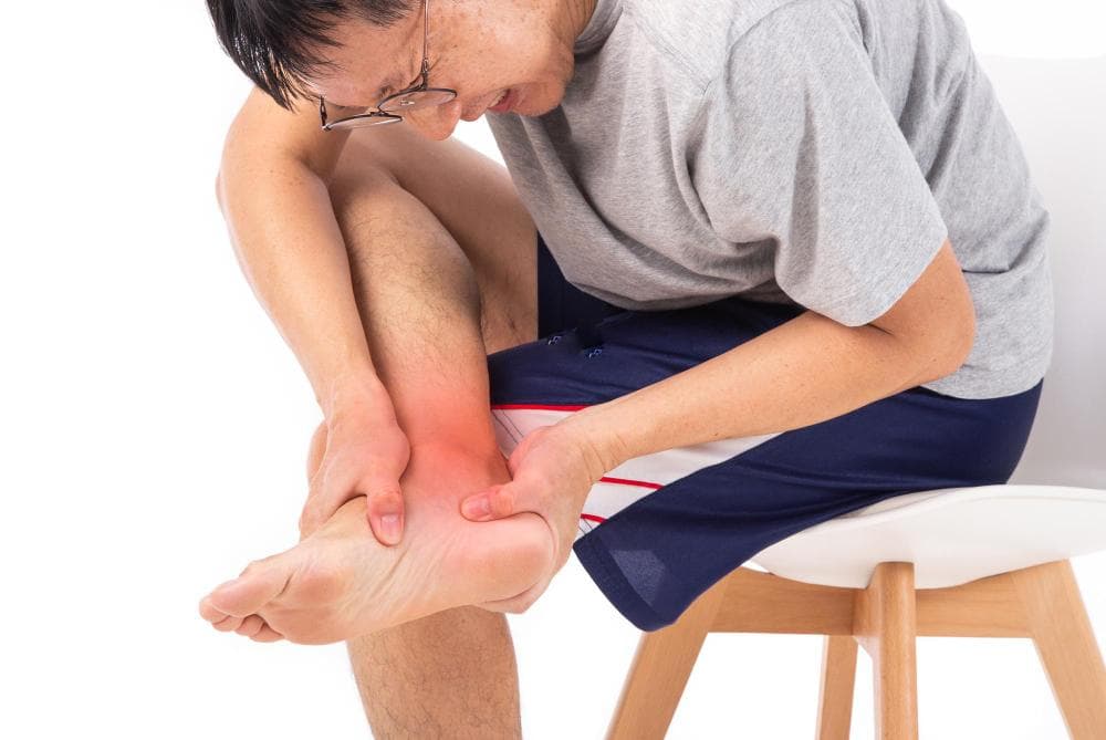 A man is sitting on a chair and holding his right ankle with an expression of discomfort. The ankle is slightly reddish in color, which is where he is holding it. The background is plain white.