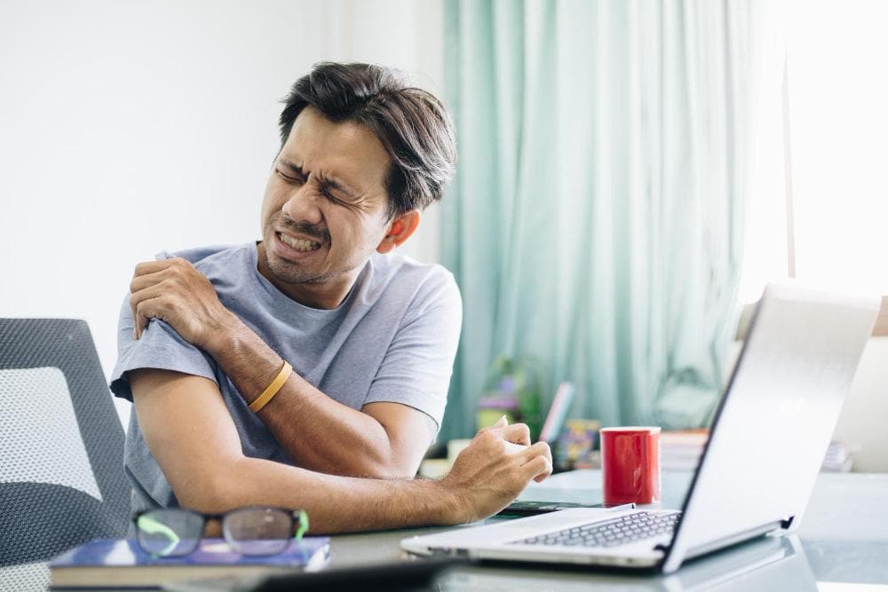 A man is sitting in front of his laptop, holding his shoulder in pain. On the table, his glasses, coffee mug, and notebook are there.