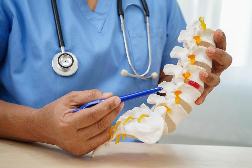 Doctor in blue scrubs holding a spine model and pointing at vertebrae with a pen to explain spinal structure and possible fusion treatment.