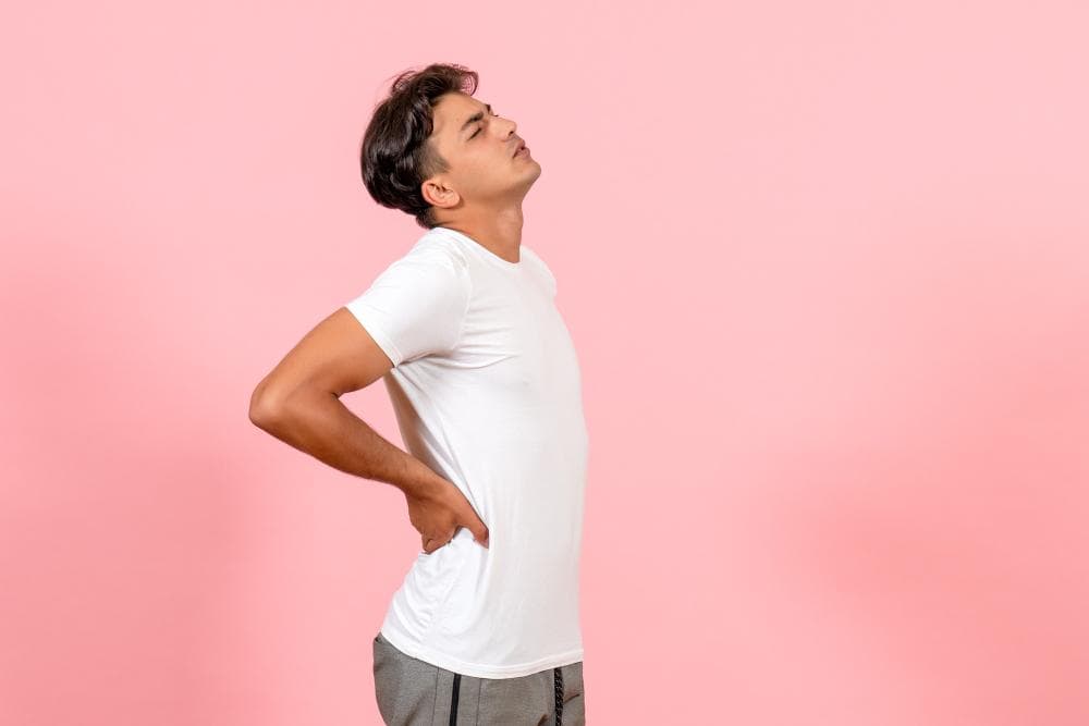 A young man stands in profile against a soft pink background, arching his back slightly while grasping his lower back with one hand. His facial expression shows discomfort as he tilts his head back.