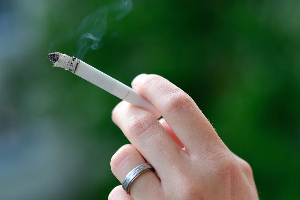 A close-up of a hand holding a lit cigarette with a small amount of ash at the tip. The person's wrist is adorned with a simple silver ring. The background is softly blurred with hints of greenery. Wisps of smoke are rising from the cigarette.