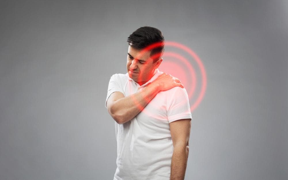 A man is seen touching his left shoulder with a concerned expression, indicating pain. The background is a soft gray, and a red graphic around the shoulder highlights areas of discomfort.