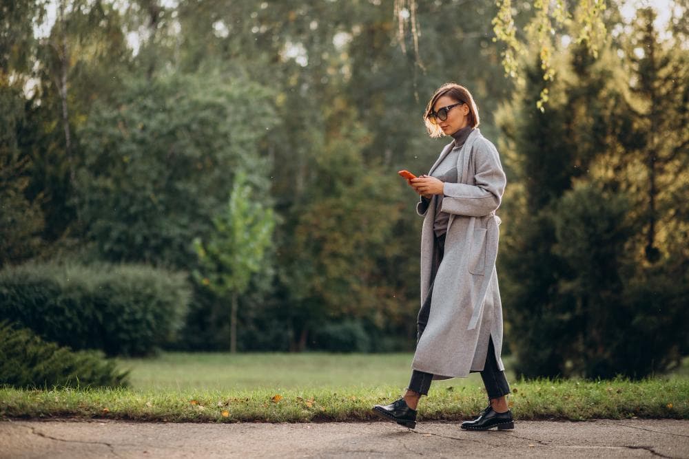 A woman walks through a park, looking at her smartphone. The background features green trees and a grassy area, with soft sunlight filtering through the leaves, creating a serene and casual atmosphere.
