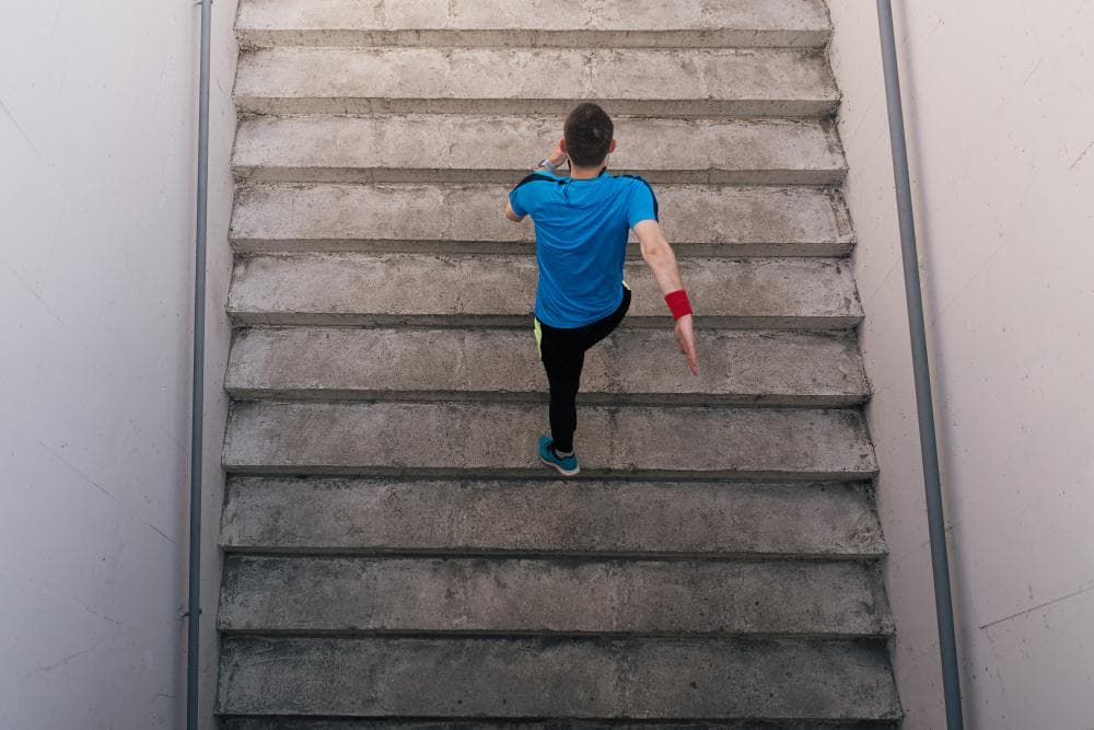 A young man is seen running up a set of concrete stairs. The perspective is from above, capturing the motion and effort as he ascends the stairs, which are well-lit and have a clean, industrial appearance.