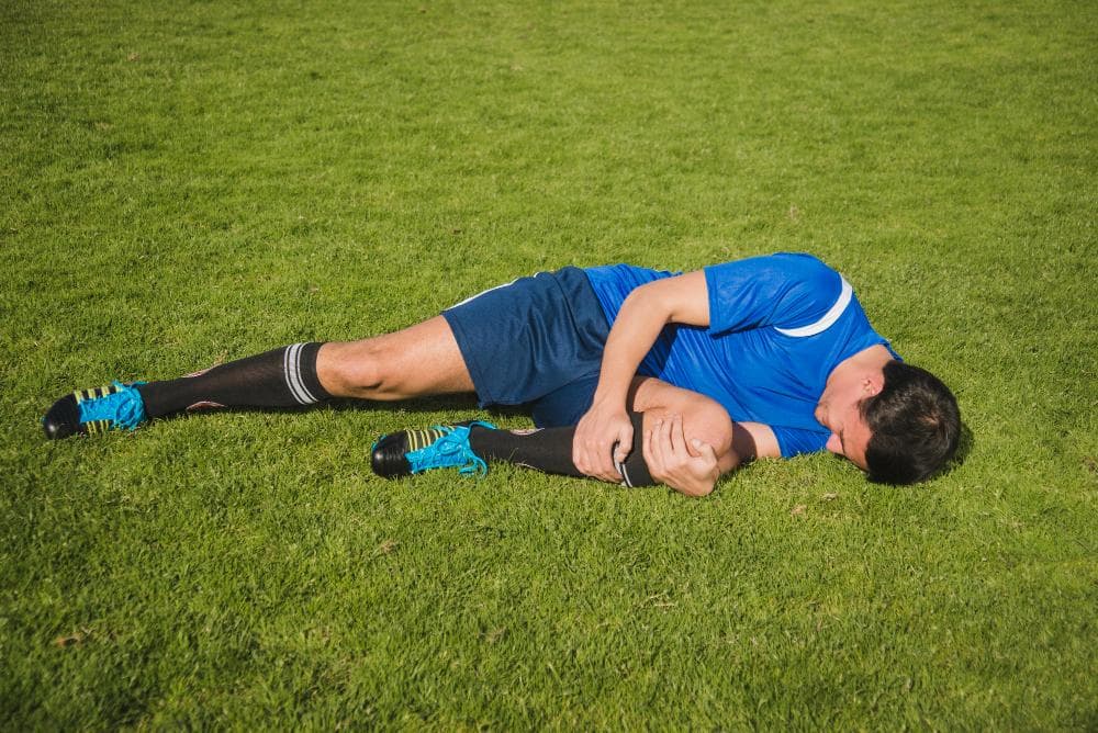 A young male soccer player in a blue jersey is lying on the grass, holding his right arm against his body as he grimaces in pain. His legs are extended on the field, and he appears to be injured, possibly after a fall or impact.