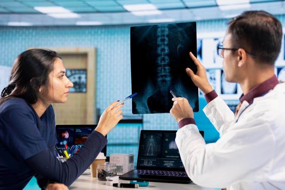 Two healthcare professionals are discussing an X-ray image showing a spine. One woman with dark hair points at the X-ray while the man in a white coat holds it up for inspection. They are surrounded by medical equipment and a laptop.