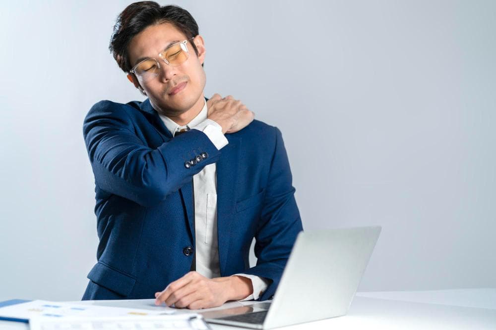 A young man in a dark blue suit is seated at a desk with a laptop, holding his left shoulder with a strained expression. His glasses add to a professional appearance, and the background is a soft gray, enhancing the focus on his discomfort.