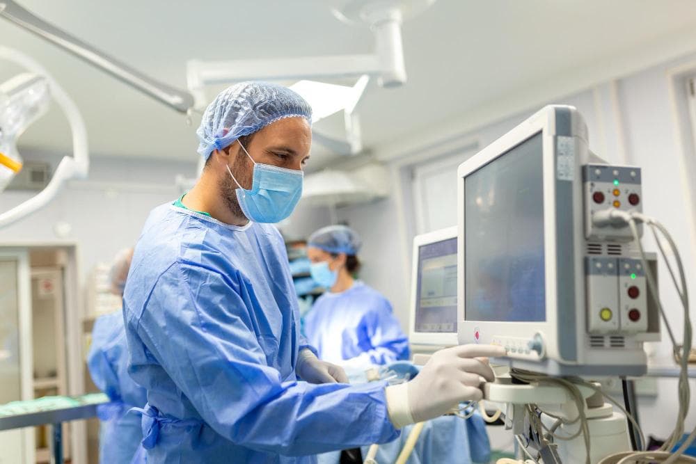 A male healthcare professional in blue scrubs is adjusting a medical monitor in an operating room. He is wearing a surgical mask and cap, with a serious expression as he focuses on the equipment. A female colleague is seen in the background.