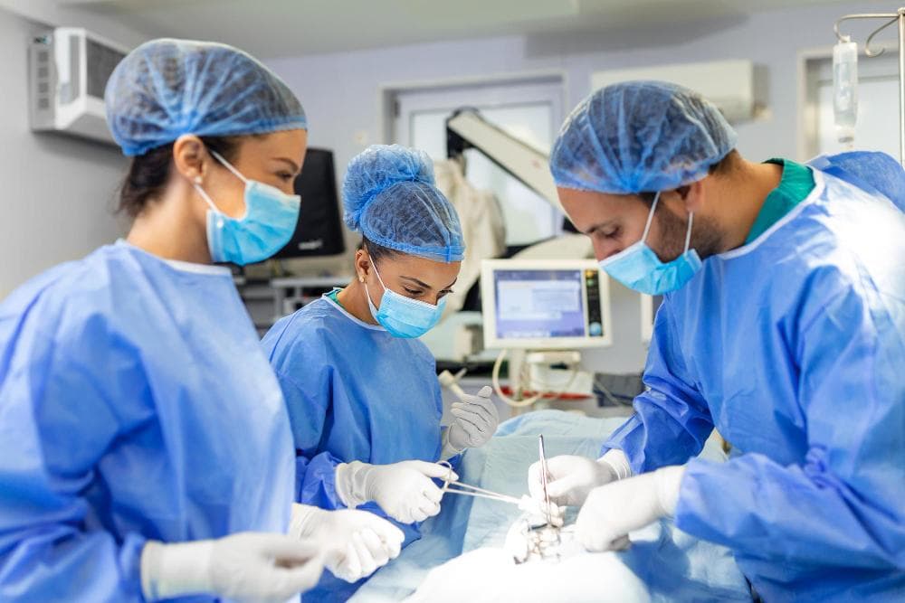 A surgical team consisting of three medical professionals is intently focused on a procedure. All are dressed in blue surgical gowns, gloves, and masks. One person is holding surgical instruments while another assists, and the third observes closely.