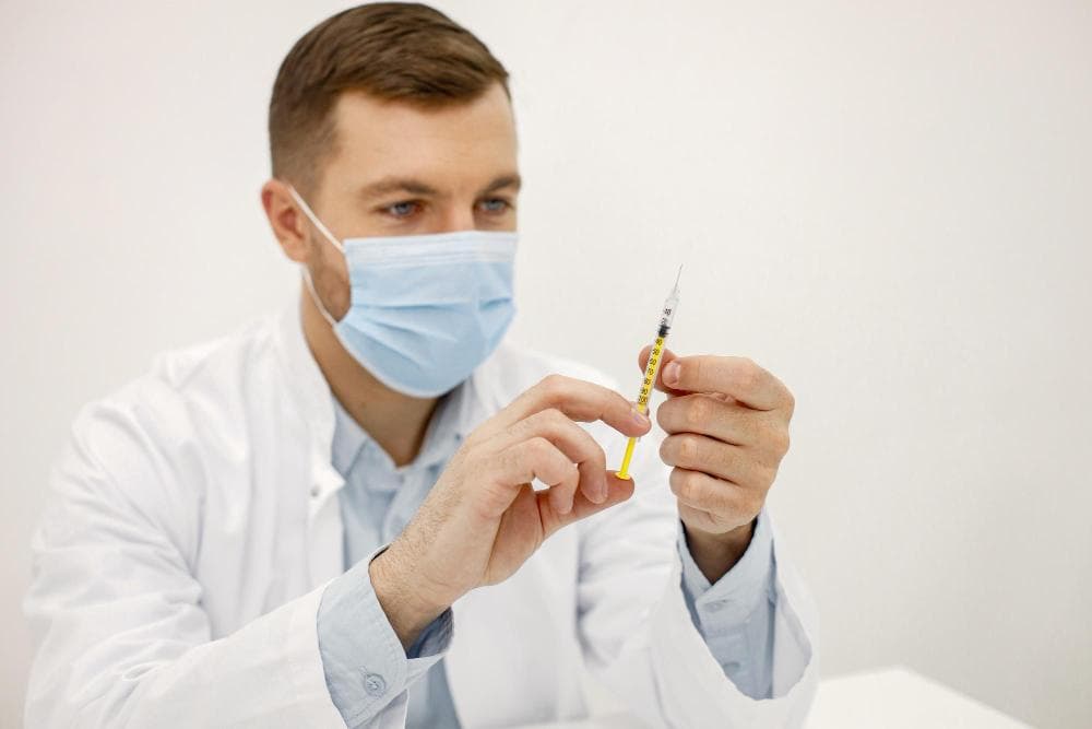 A male doctor in a white coat and blue mask is examining a syringe filled with medication, holding it up for closer inspection. His focused expression suggests he is checking the dosage or preparing for a procedure.